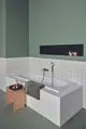 Badezimmer mit freistehender Badewanne, weißen Fliesen und grünem Anstrich. Holztablett mit Produkten, darunter eine Handbrause von Ideal Standard. Handtuch auf der Badewanne.
